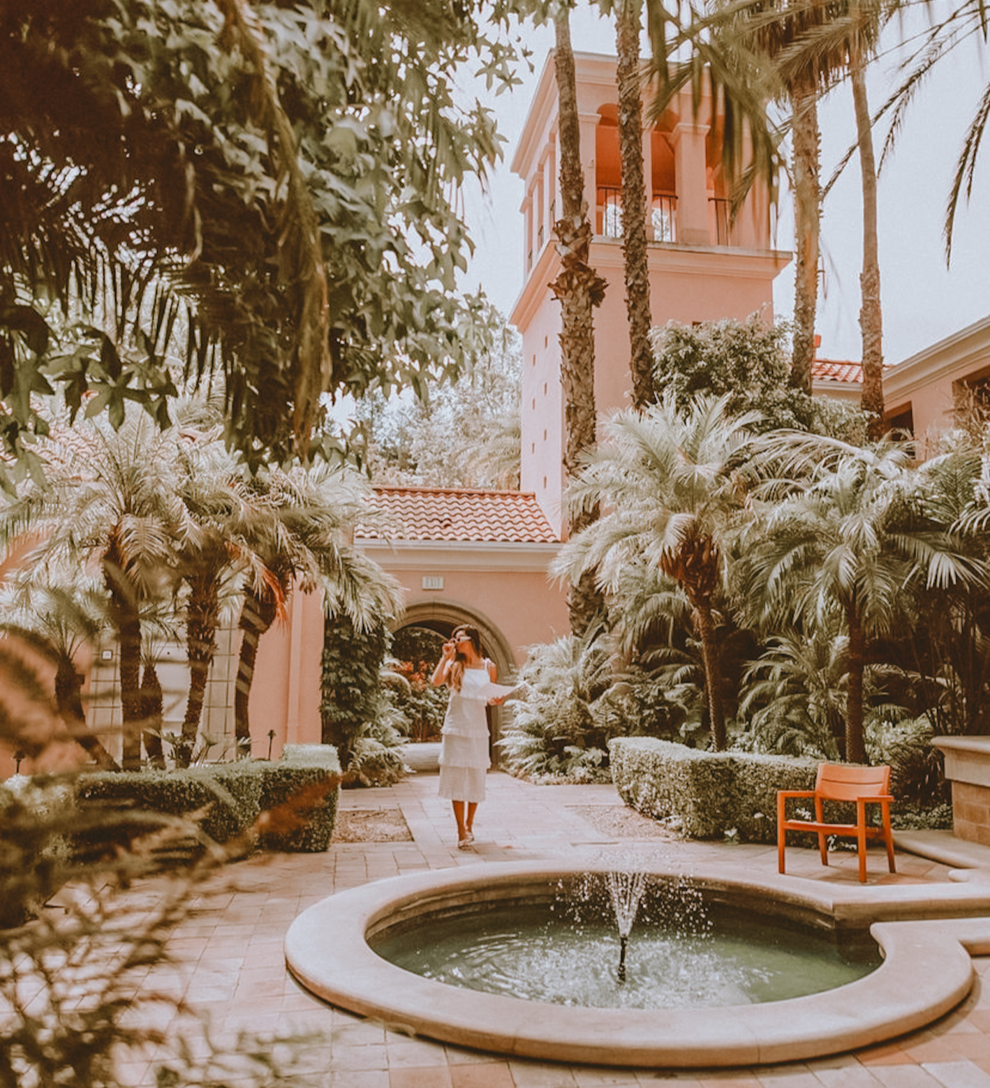 A tranquil courtyard featuring a circular fountain, lush palm trees, and a pink stucco building with arched windows and a tower. A woman in a white dress stands near the fountain.