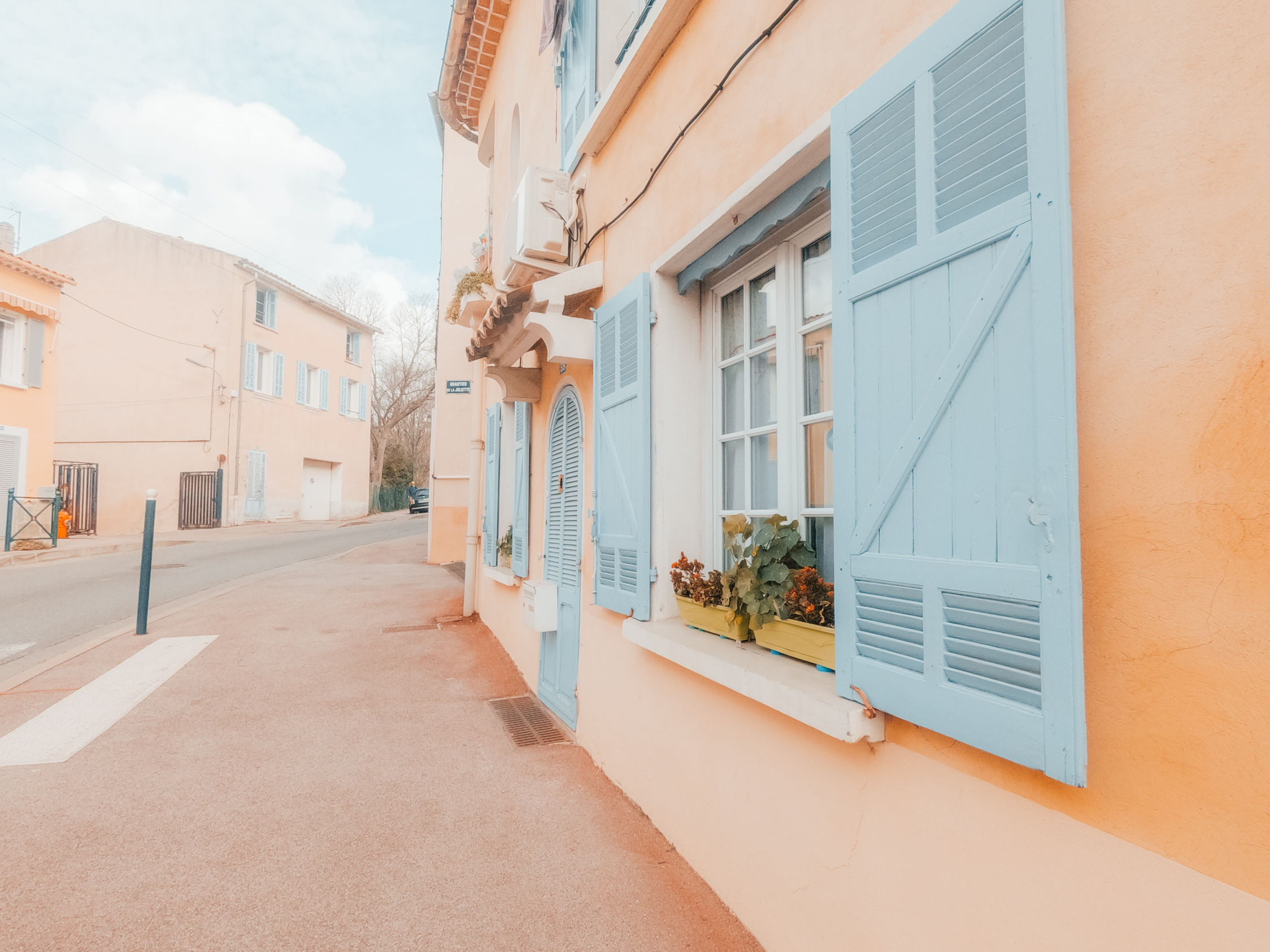 A charming street with pastel peach buildings featuring bright blue shutters and window boxes filled with flowers, under a clear blue sky.