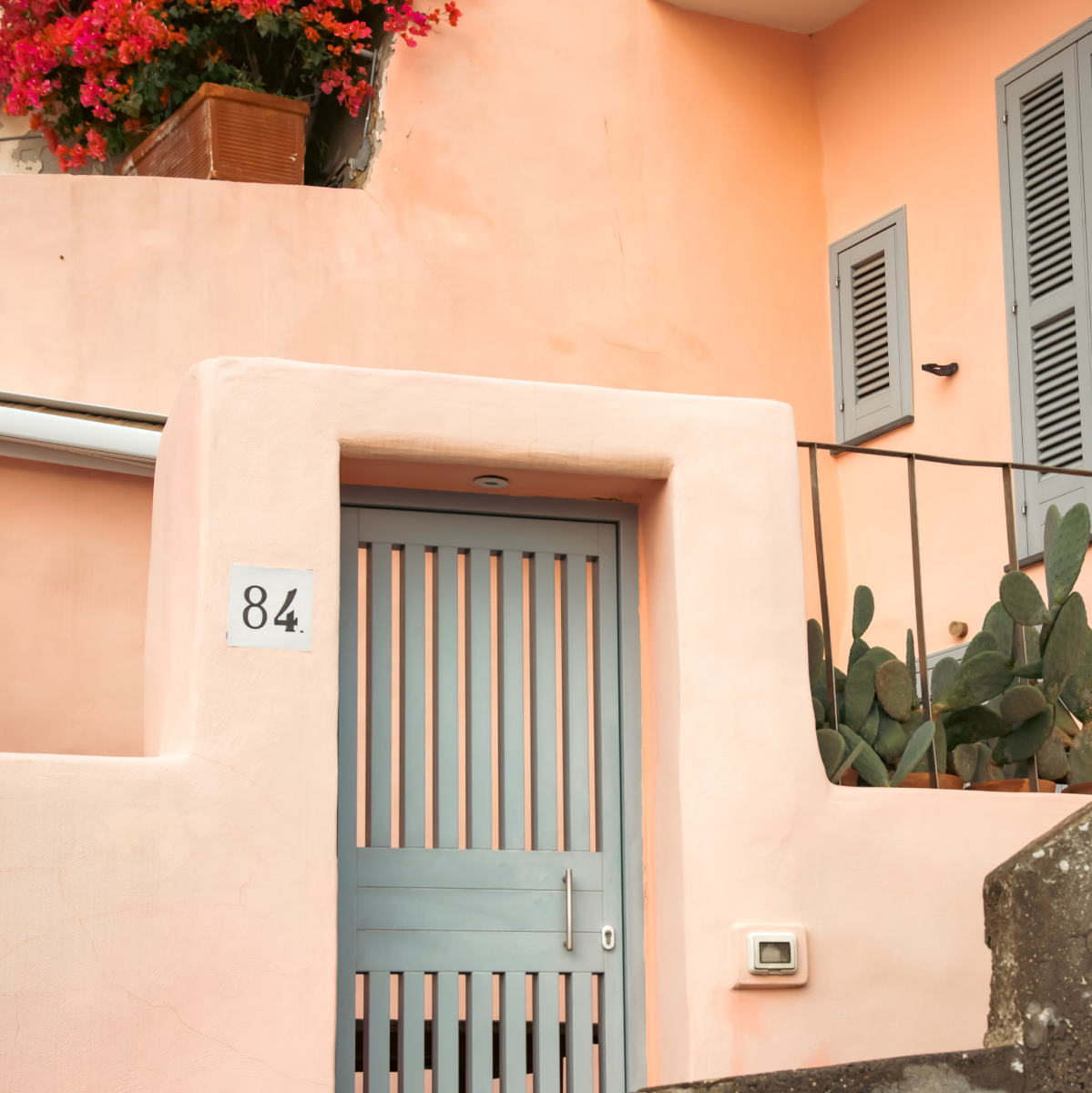 A pastel peach house featuring a modern gray gate, potted cactus plants, and vibrant pink bougainvillea flowers on a balcony.