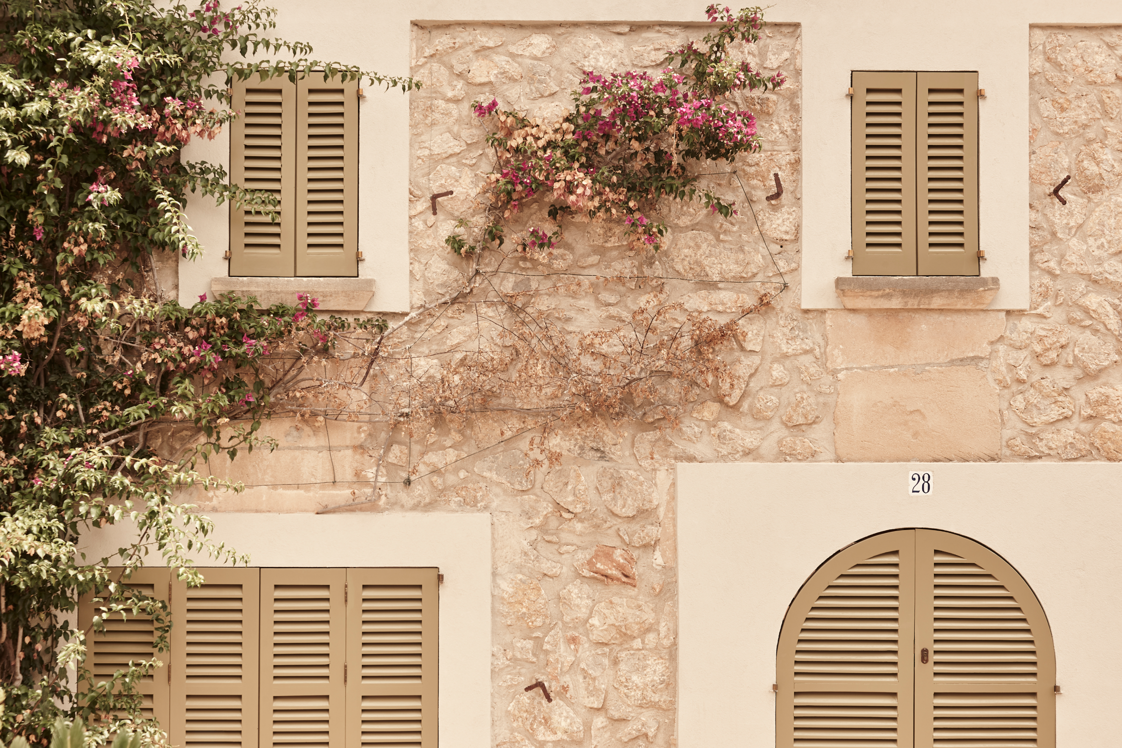 A rustic stone facade with beige shutters on windows and an arched door, adorned with climbing bougainvillea in vibrant pink blooms.