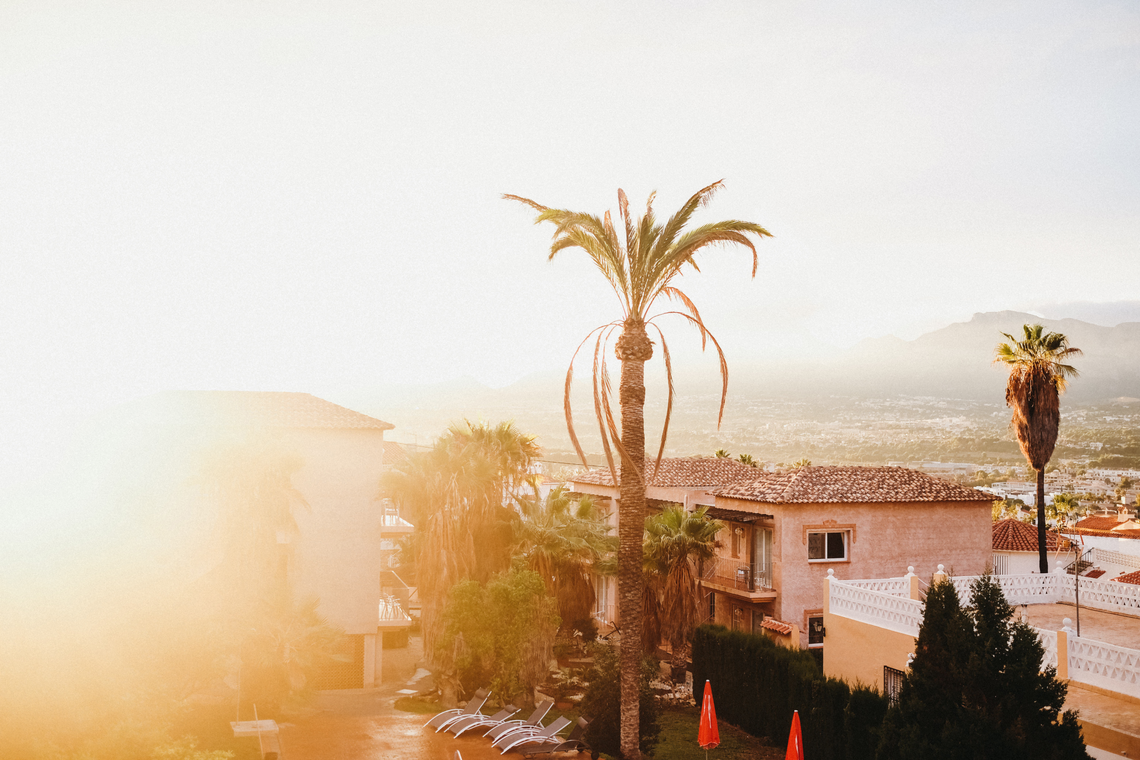 A scenic view of a Mediterranean-style villa surrounded by palm trees at sunset, with mountains and a hazy golden sky in the background.